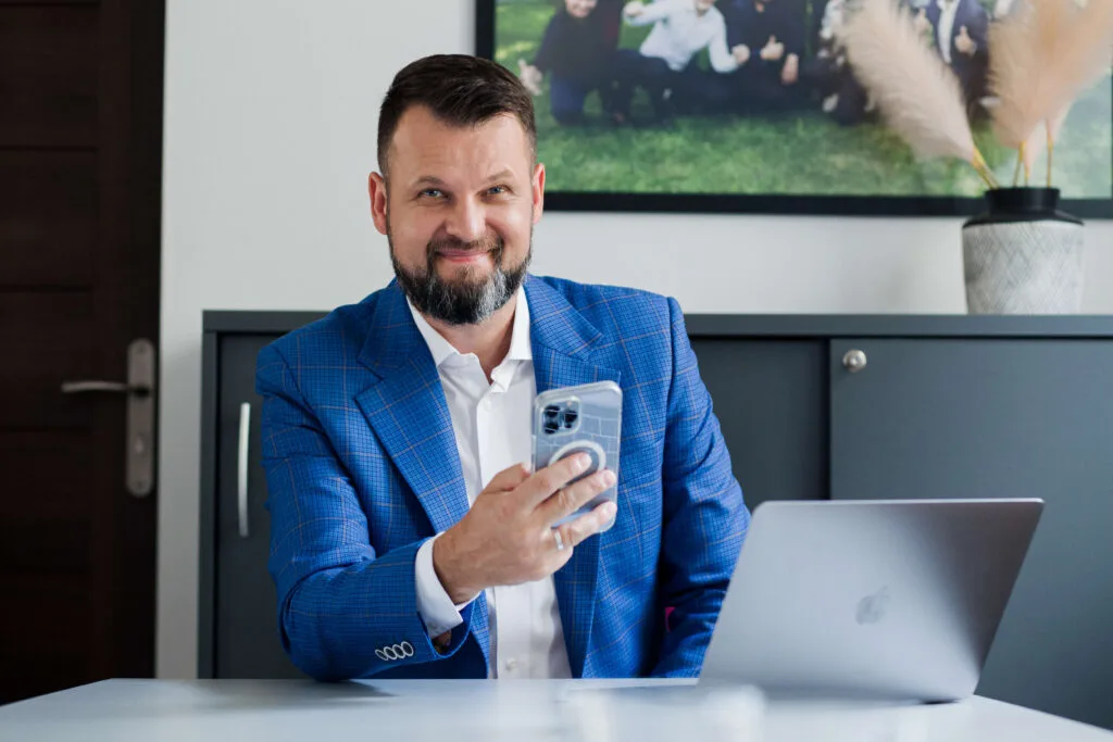Zdjęcie uśmiechniętego mężczyzny w niebieskim garniturze siedzącego przy stole z telefonem komórkowym w ręce.