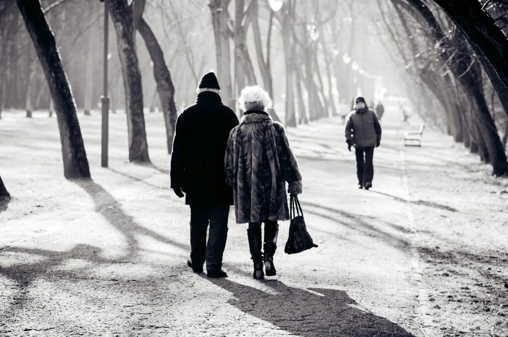 Czarno-biała fotografia przedstawiająca parę seniorów idących aleją parkową.