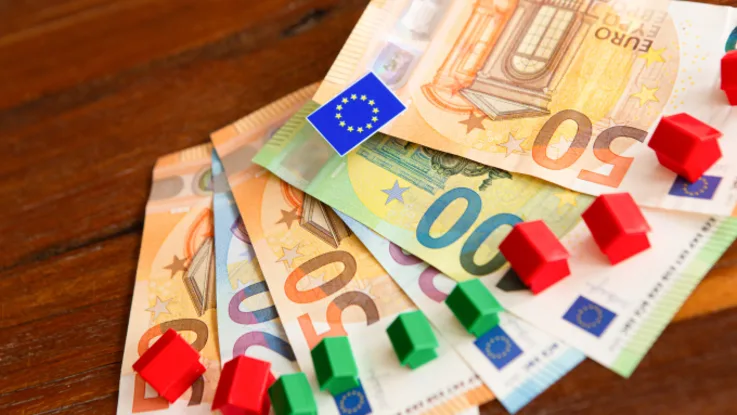 Zdjęcie banknotów waluty EURO oraz stojących na nich miniaturowych domków w kolorze zielonym i czerwonym.
