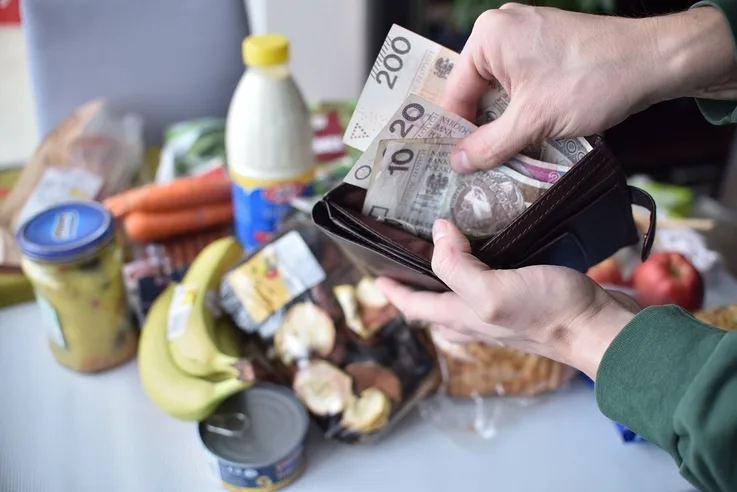 Zdjęcie dłoni wyciągającej banknoty z portfela na tle produktów spożywczych.